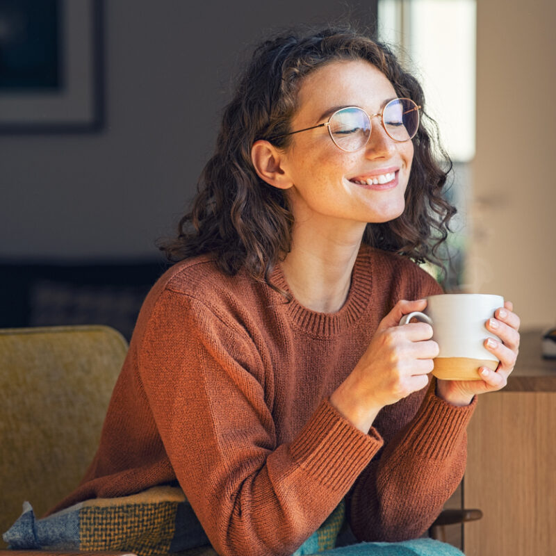 Fall HVAC Tips for Tampa Homes: Stay Comfortable as Temperatures Change, Happy young woman drinking a cup of tea in an autumn morning. Dreaming girl sitting in living room with cup of hot coffee enjoying under blanket with closed eyes. Pretty woman wearing sweater at home and enjoy a ray of sunshine on a winter afternoon.